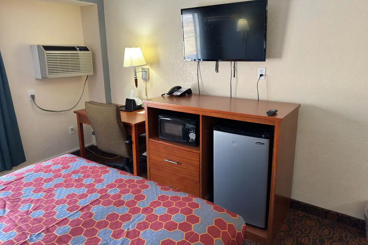 A hotel room with a bed, a desk with a microwave, a refrigerator, a television, and an air conditioning unit.