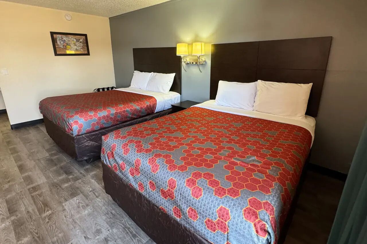 Two beds with matching red and blue patterned bedspreads, a dark headboard, a wall-mounted light fixture, and a framed picture on the wall.
