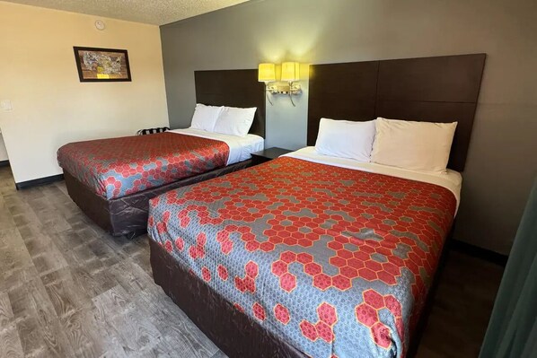 Two beds with matching red and blue patterned bedspreads, a dark headboard, a wall-mounted light fixture, and a framed picture on the wall. - Ember Inn Suites Waite Park (Waite Park)