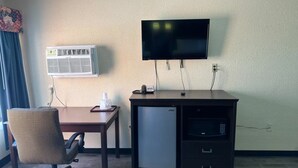Una habitación con escritorio, silla, televisor, aire acondicionado y un refrigerador pequeño.