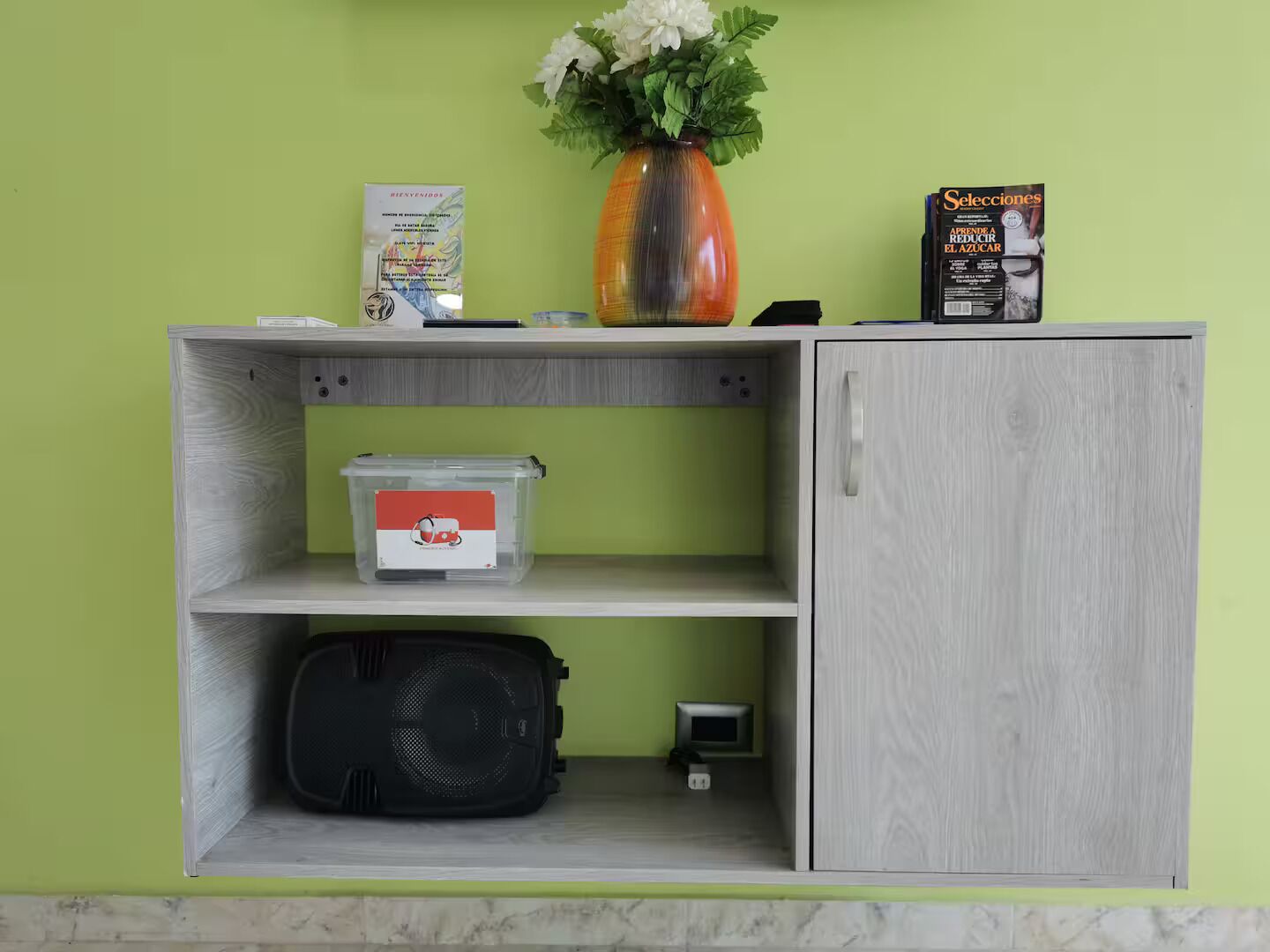 Ein kleiner, heller Holzkabinent mit einer Ablage, einer Vase mit weißen Blumen, einer Zeitschrift und einem kleinen elektronischen Gerät.