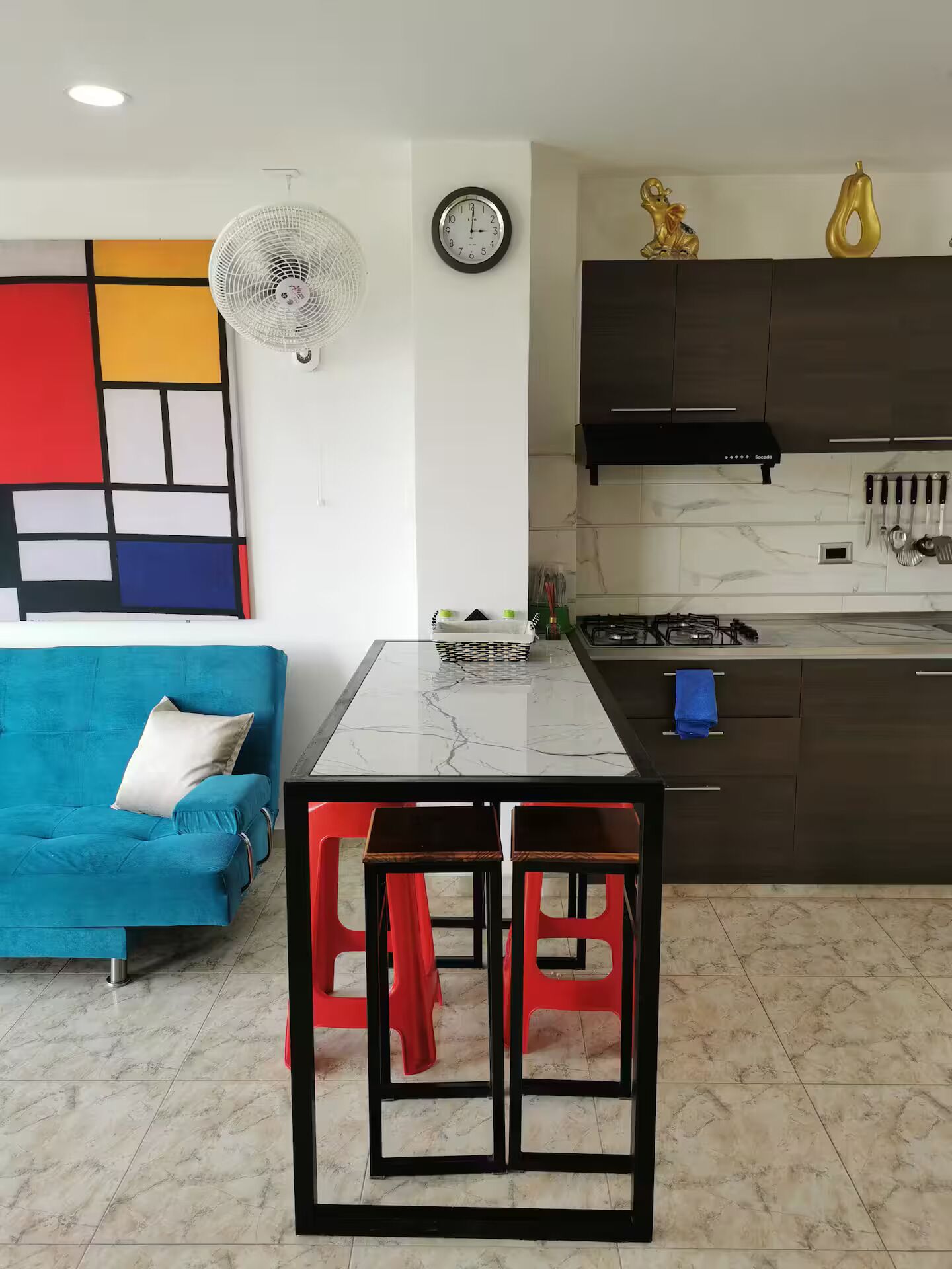 Een moderne keuken met een marmeren aanrecht, een eettafel met rode krukken, een blauwe bank en een wandklok.