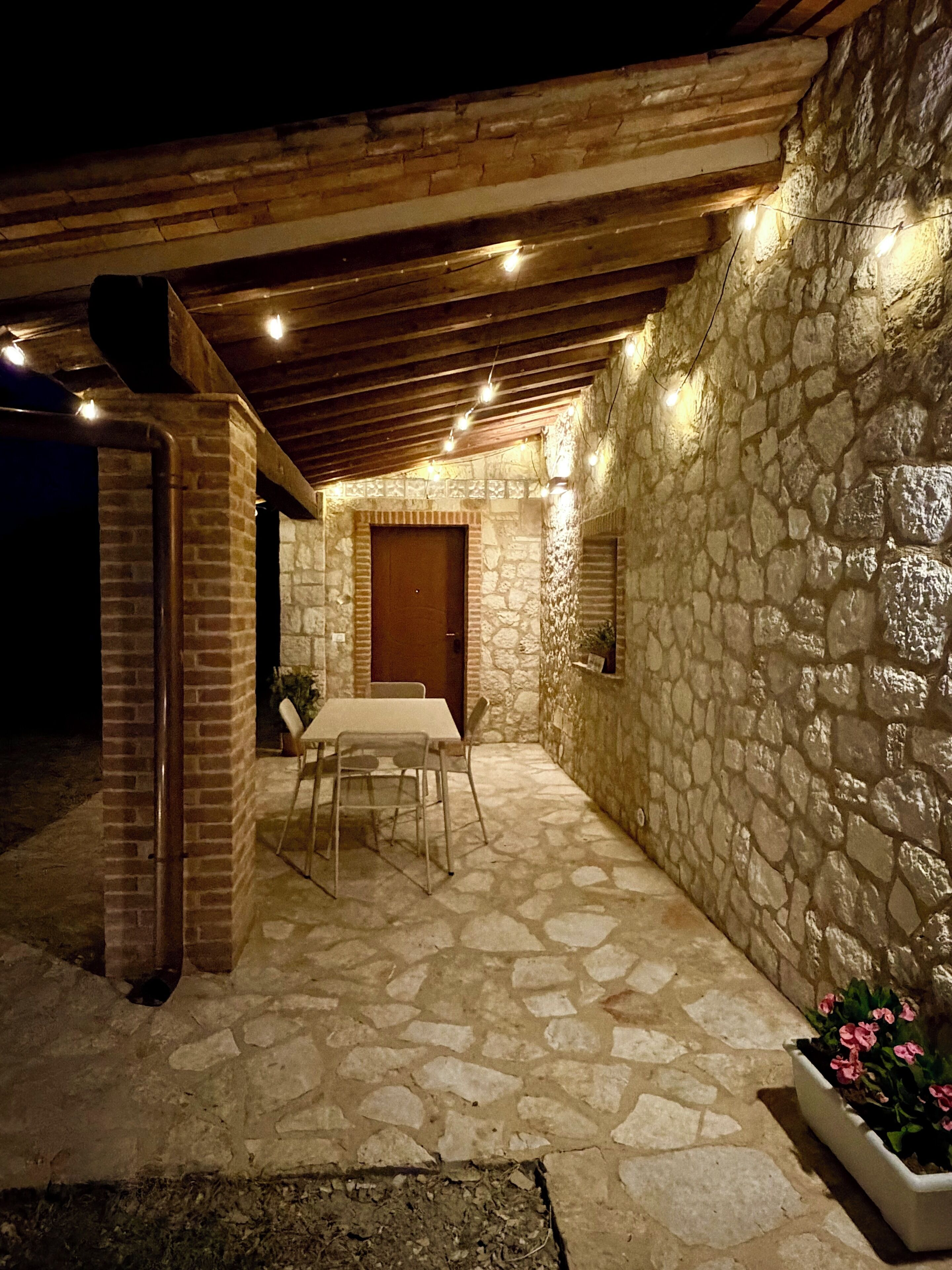 Une terrasse extérieure avec des murs en pierre, une table et des chaises, un plafond en bois et des guirlandes lumineuses.