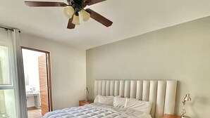 A bedroom with a bed, wooden floor, ceiling fan, bedside tables, and a view through a door.