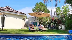 A swimming pool with two lounge chairs and an umbrella, surrounded by greenery and a building with a tiled roof.