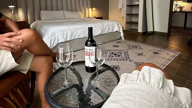 A hotel room with a bed, a ceiling fan, a table with a bottle and glasses, and a patterned rug.