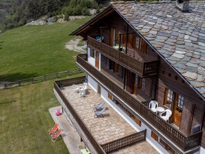 A two-story wooden chalet with a stone roof, a balcony with outdoor seating, and a view of a green landscape. - Chalet Remondet by Interhome (Sarre)