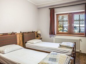 A room with two single beds, a wooden headboard, a nightstand with a lamp, a window with curtains, and a radiator. - Chalet Remondet by Interhome (Sarre)
