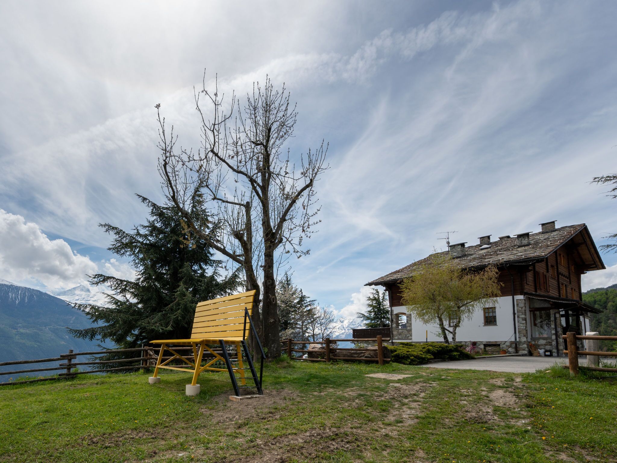 A wooden cabin with a stone foundation, a wooden fence, and a bench in a grassy area with a mountain backdrop.
