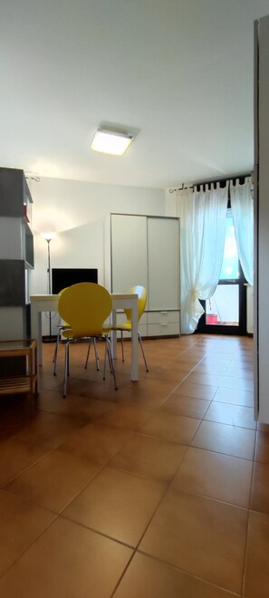 A room with a tiled floor, a yellow chair, a white table, and a window with white curtains. - Piazzale Unità d'Italia 3B (Udine)