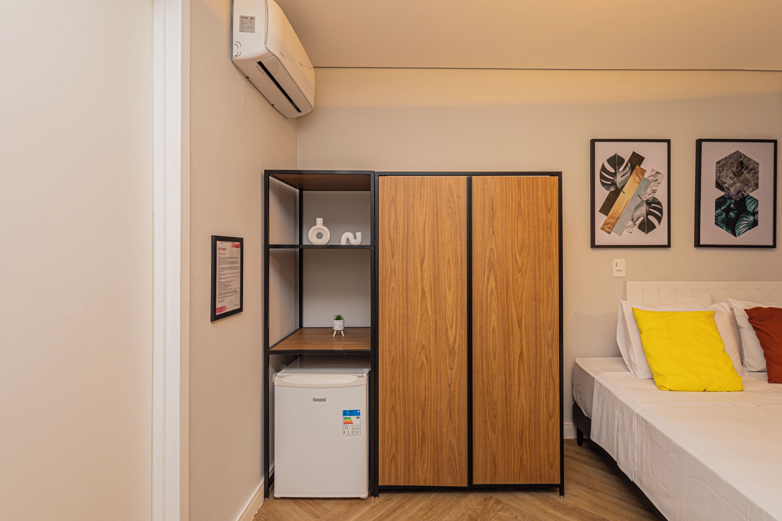Een slaapkamer met een bed, een kledingkast, een kleine koelkast en een luchtverkoeler aan de muur.