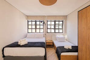 Habitación de hotel con dos camas, una mesita de noche, una lámpara y ventanas amplias con vistas a edificios.