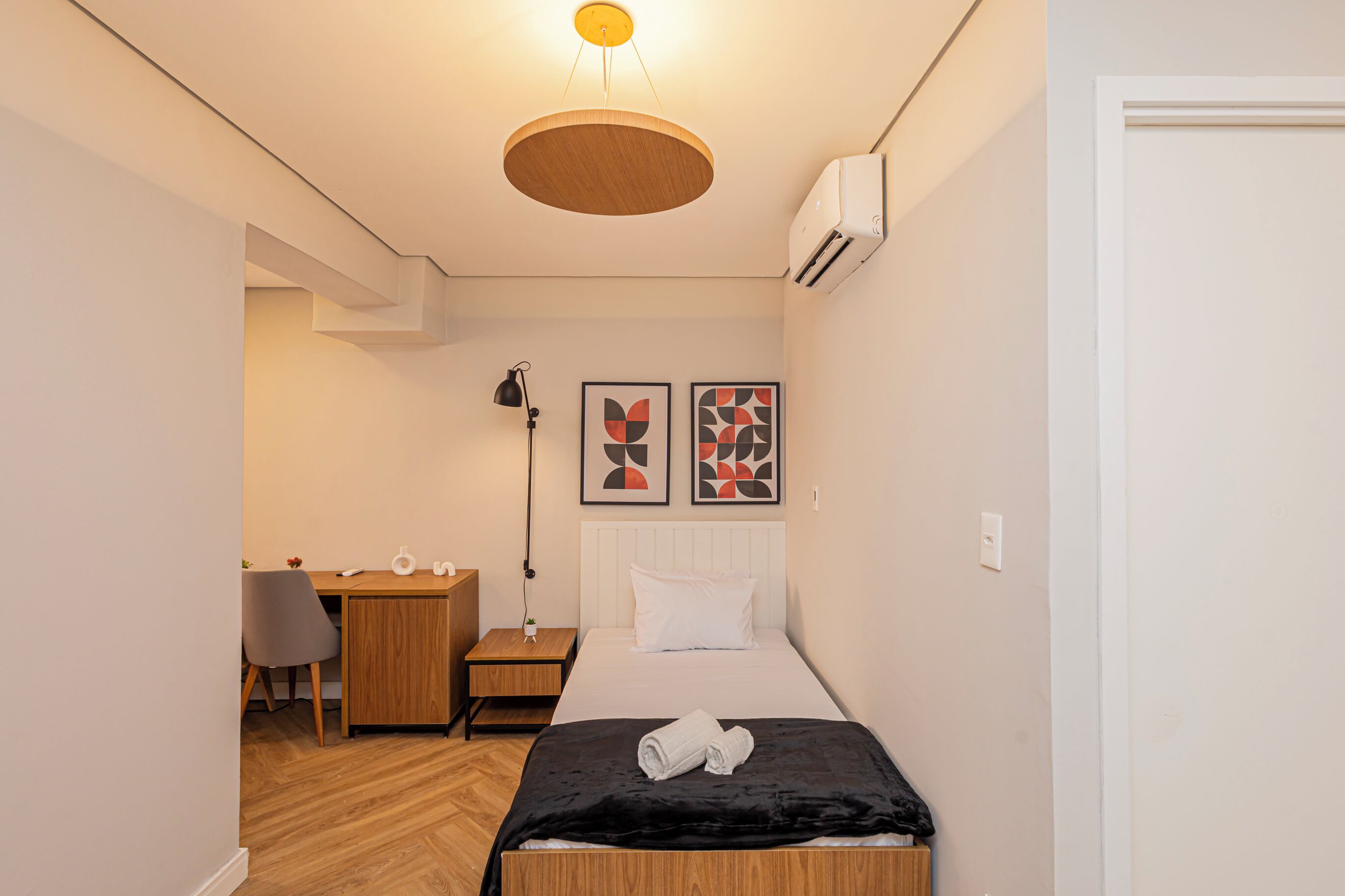 A compact hotel room with a bed, a desk, a chair, a lamp, and two abstract wall art pieces.