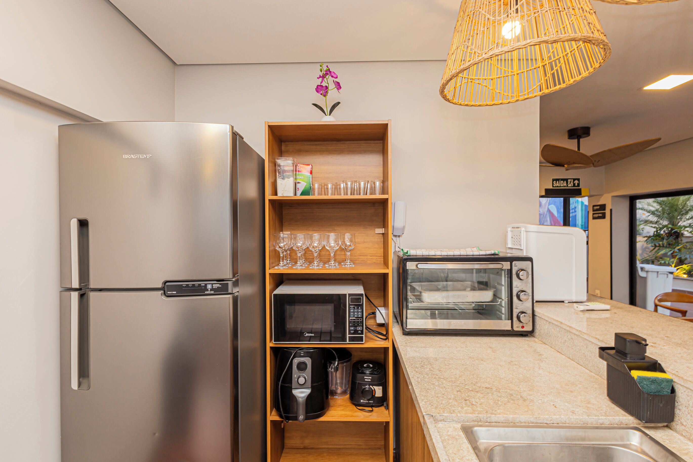 A compact kitchen with a stainless steel refrigerator, a microwave, a toaster, and a shelf with glassware and a coffee maker.