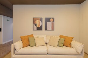 A two-seater sofa with multiple cushions, a framed abstract artwork above it, and a door to the left. - One House by Anora Spaces (São Paulo)