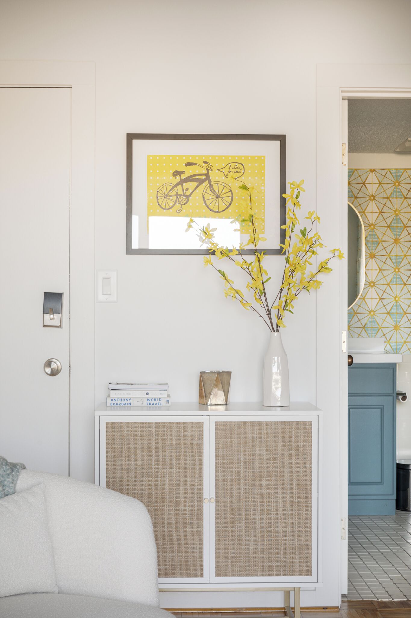 Una stanza con una porta bianca, un quadro con una bicicletta, un vaso con fiori gialli, un piccolo armadietto e uno specchio con motivo geometrico.