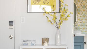 Una stanza con una porta bianca, un quadro con una bicicletta, un vaso con fiori gialli, un piccolo armadietto e uno specchio con motivo geometrico.