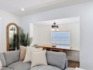 Ein Wohnzimmer mit grauem Sofa, einem Holzesstisch, einem Spiegel und einem Fenster mit Jalousien.