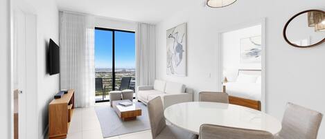 Una habitación de hotel moderna con comedor, sofá, televisor y un amplio ventanal con vista a la ciudad.