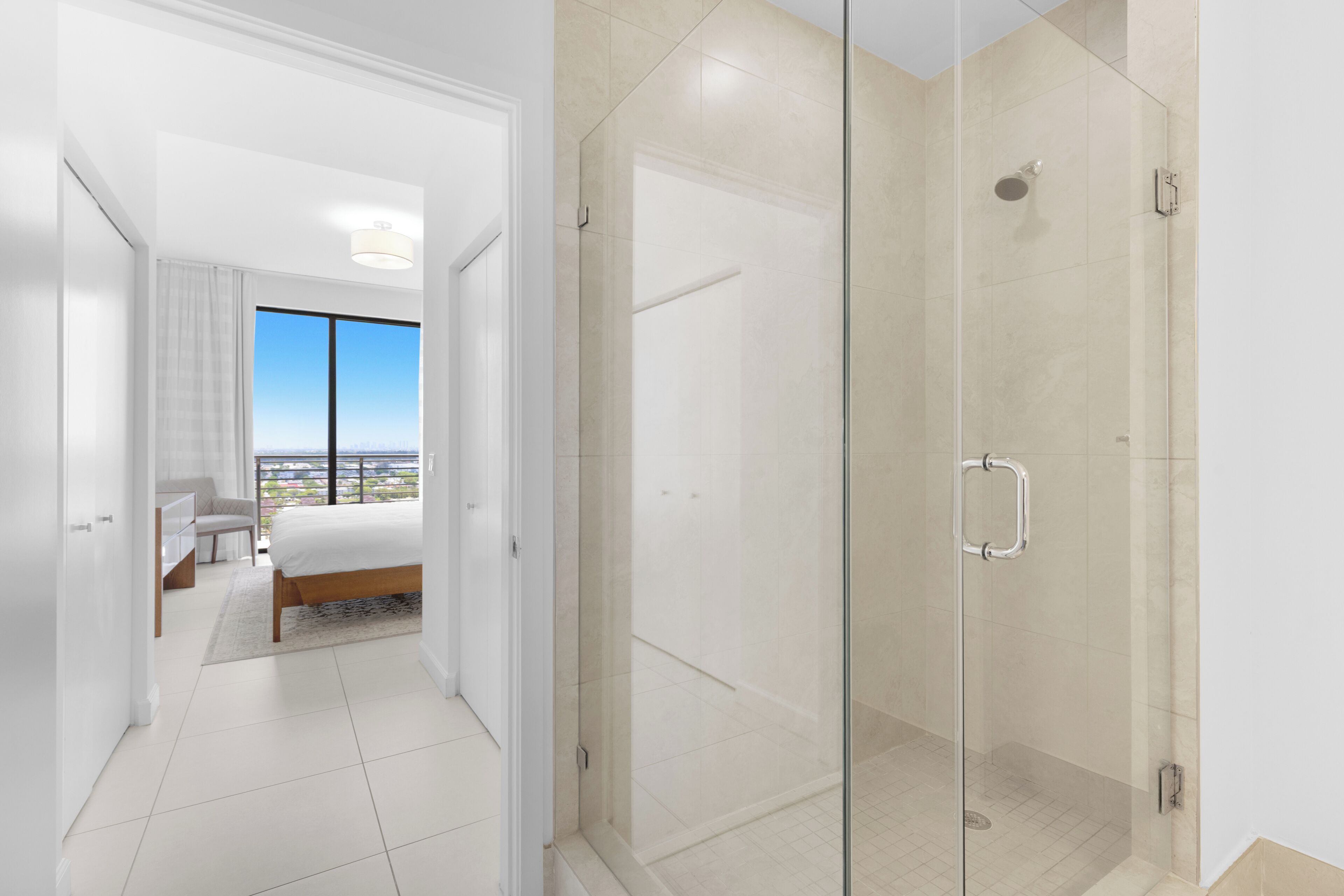 A modern hotel room with a glass-enclosed shower, a bed, a desk, and a view of the city.