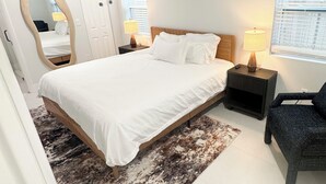 Une chambre à coucher comprenant un lit, deux tables de chevet avec des lampes, un ventilateur de plafond, un grand miroir et une fenêtre avec des stores.