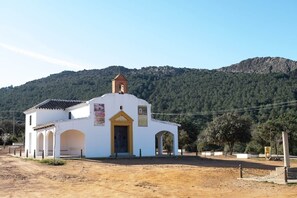A small white building with a bell tower, surrounded by a dirt area and a forested hill in the background. - El Puente Grande Country House · Pool and Garden (Navahermosa)