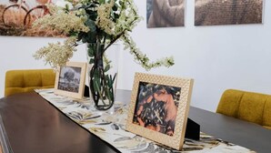 Ein Esstisch mit Blumen in einer Vase, gerahmten Bildern und einer gemusterten Tischläufer.