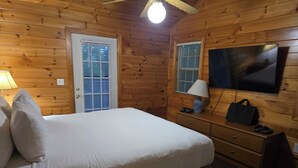 Ein Zimmer in einer Holzhütte mit einem Bett, einem Schminktisch, einem Fernseher und einer Tür mit Jalousien.