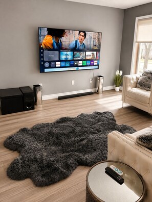 Ein Wohnzimmer mit flachbildfernseher an der Wand, einem flauschigen Teppich, einer Couch, einem Couchtisch und einem Lautsprecher.