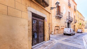 A narrow cobblestone street with buildings featuring traditional architecture, a metal gate, and parked vehicles.