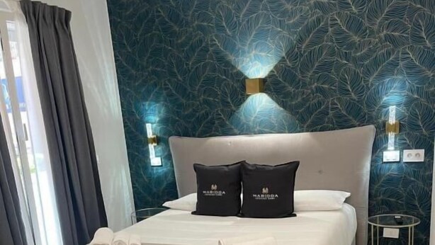Una habitación de hotel con una cama, una mesa de madera, una silla color turquesa, una mesita de noche y una pared con un diseño de hojas.