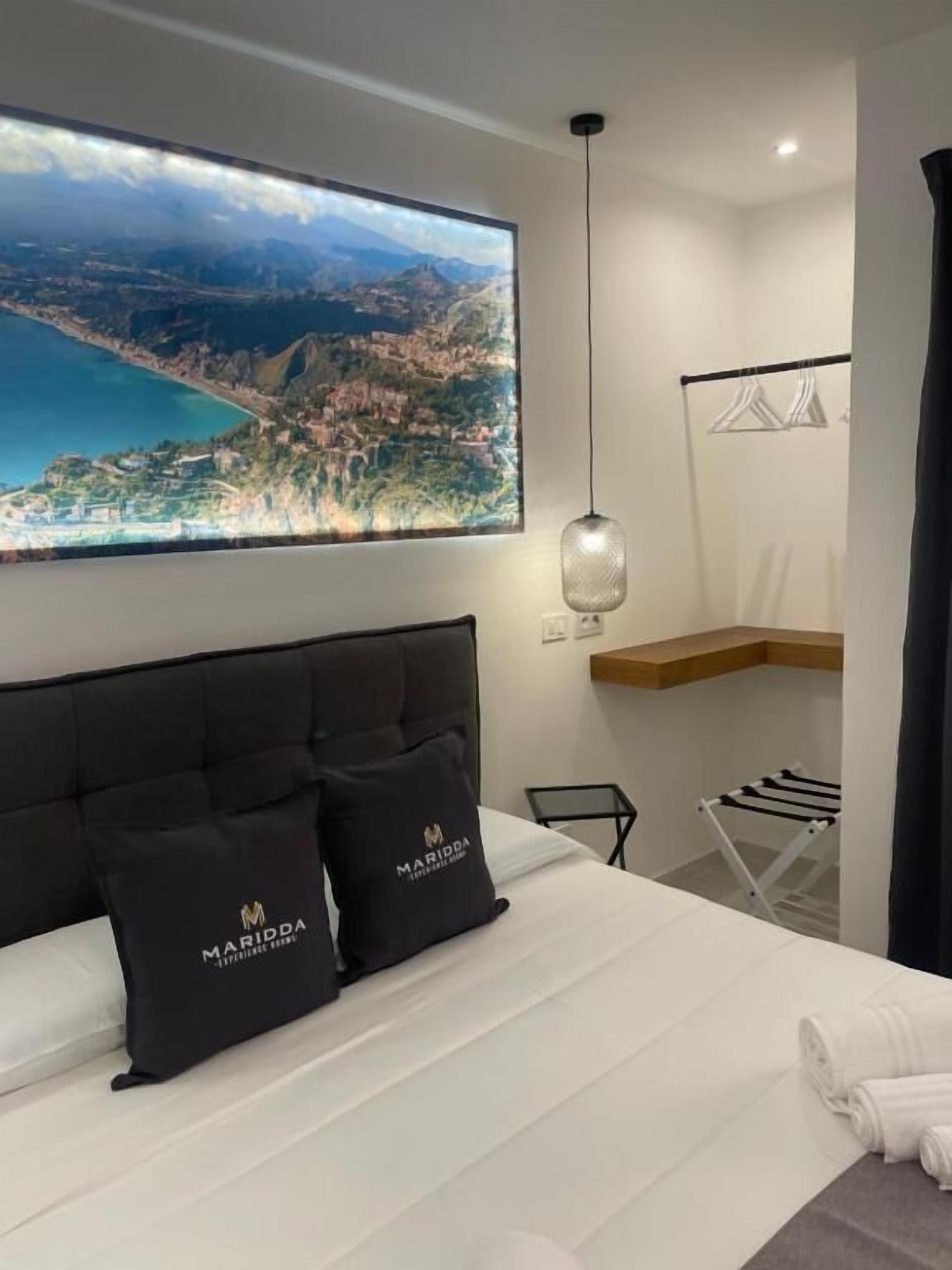 Une chambre d’hôtel avec une grande fenêtre, un lit avec deux oreillers, un luminaire suspendu, une étagère en bois et un étendoir.
