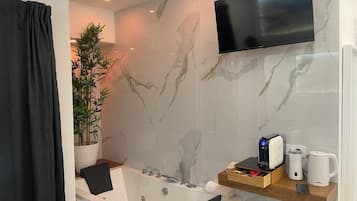 A modern bathroom with a marble wall, a mounted TV, a bathtub, a plant, and a coffee maker.