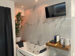 A modern bathroom with a marble wall, a mounted TV, a bathtub, a plant, and a coffee maker.