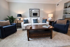 A living room with a white sofa, two blue armchairs, a wooden coffee table, and a large mirror on the wall.