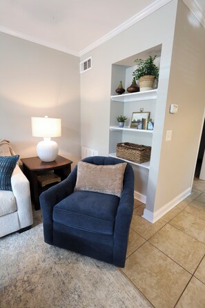 A living room with a blue armchair, a white lamp, a small table, and a white shelving unit with decorative items.
