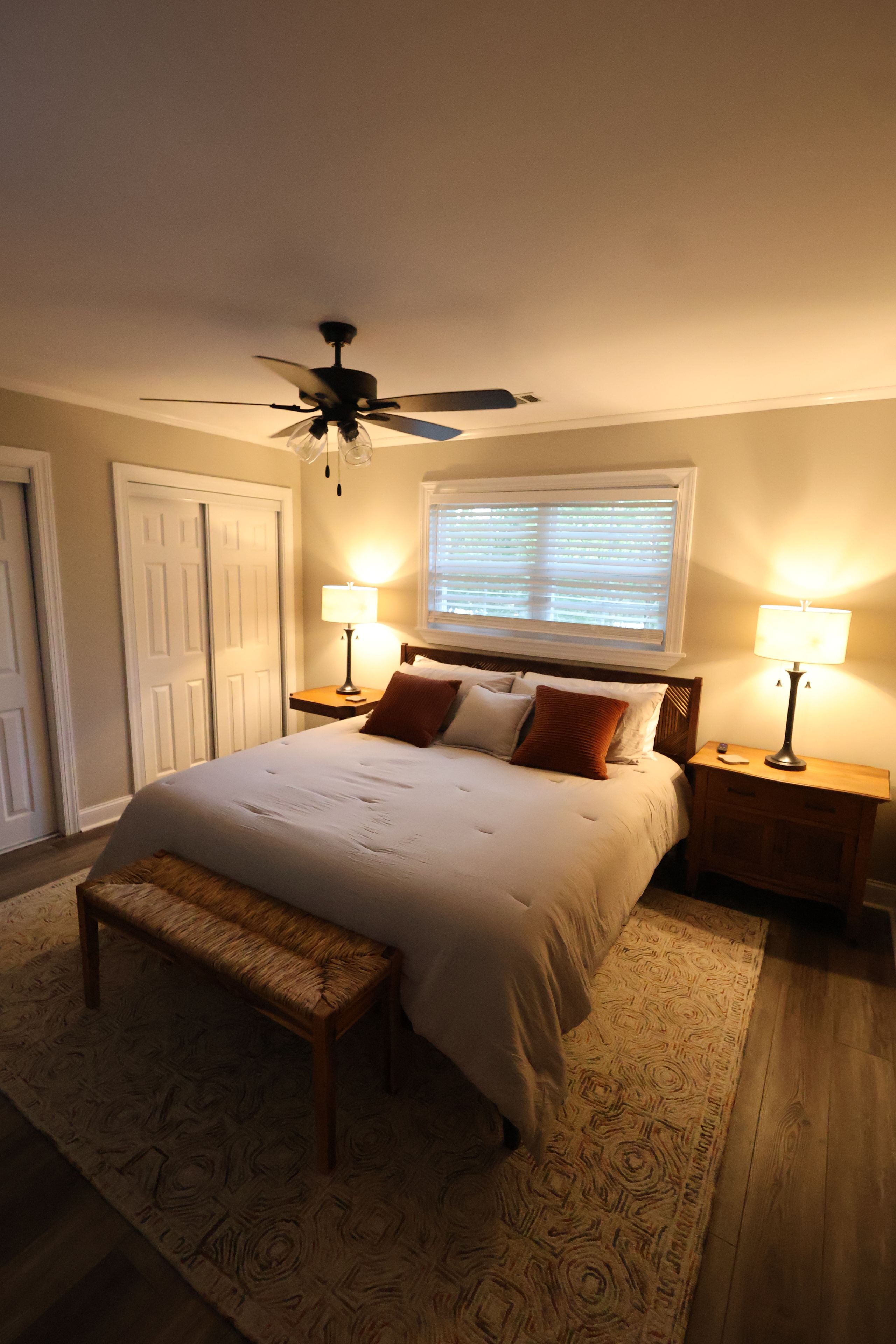 A bedroom with a bed, two bedside tables with lamps, a ceiling fan, a window with blinds, and a door.