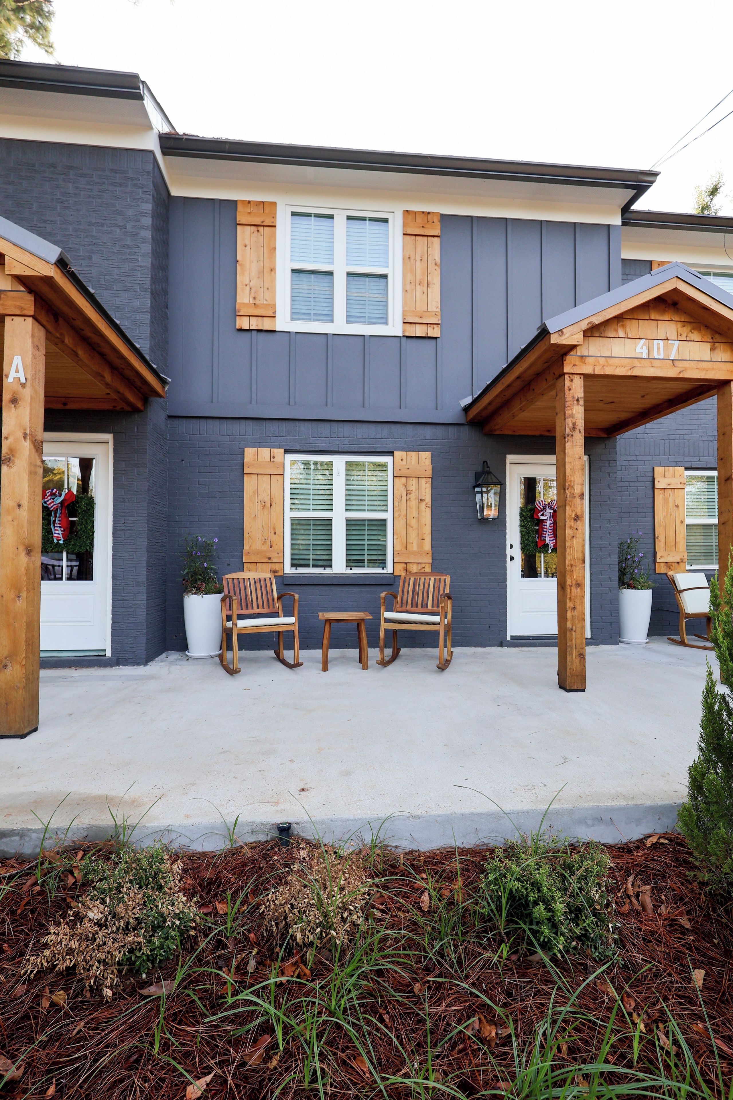 A two-story building with a grey facade, wooden shutters, and a covered porch area with two wooden chairs and a small table.