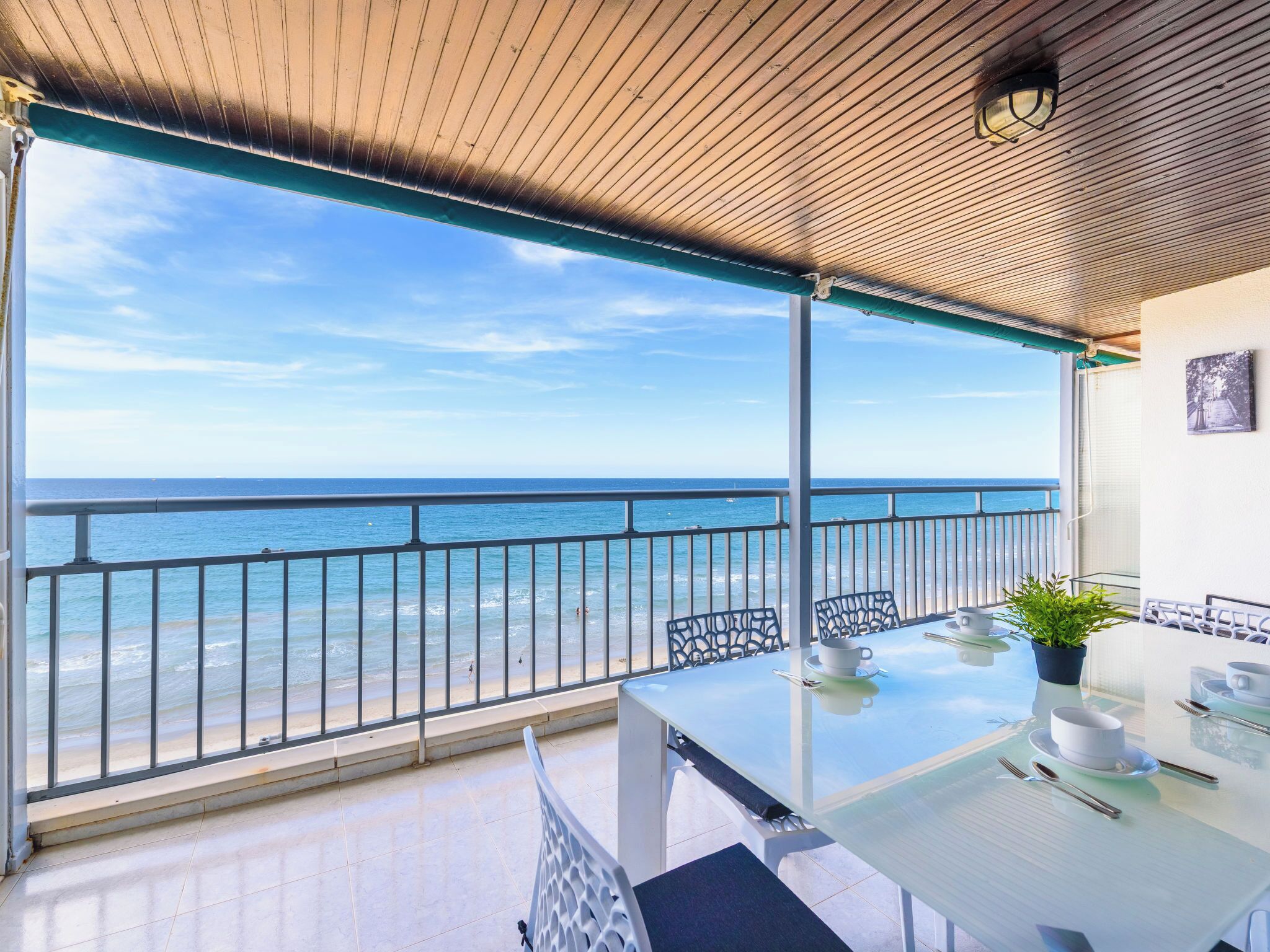 A balcony with a glass table, chairs, a potted plant, and a view of the sea.