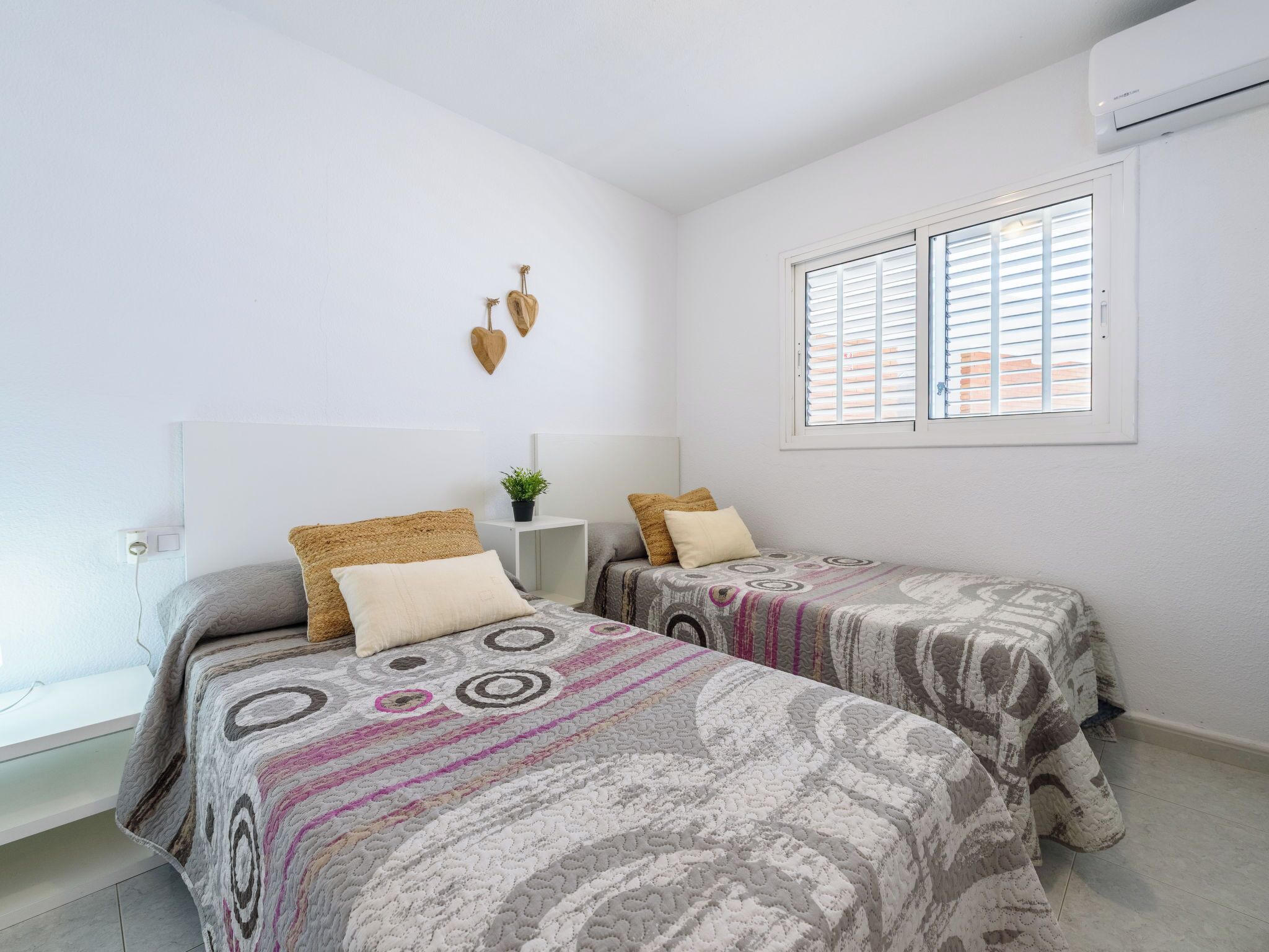 A bedroom with two beds, a small bedside table with a plant, a wall-mounted air conditioner, and a window with blinds.