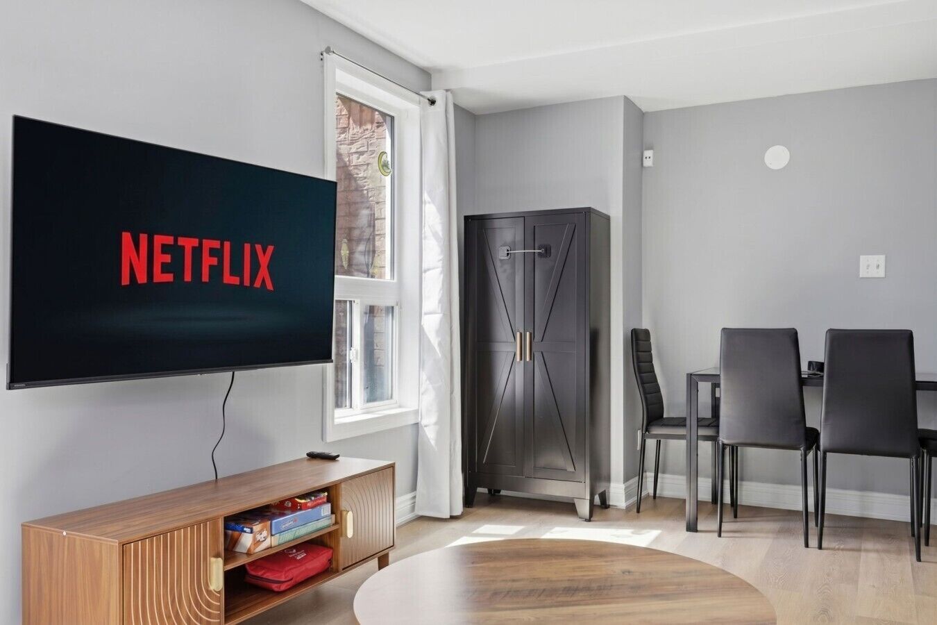 A modern living room with a flat-screen TV displaying 'NETFLIX,' a wooden entertainment unit, a black refrigerator, and a dining area with two chairs.