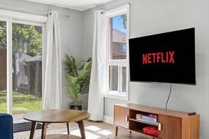 Olohuoneessa on suuri televisio, jossa näkyy Netflix, puinen pöytä, ruukkukasvi ja suuret ikkunat, joista avautuu näkymä ulos.