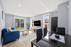 A modern living room with a blue sofa, a dining table with chairs, a TV displaying 'NETFLIX,' and a sliding glass door leading to a garden. - Private 1BR Apt | WiFi | Parking | Nr all Amenities (Mississauga)