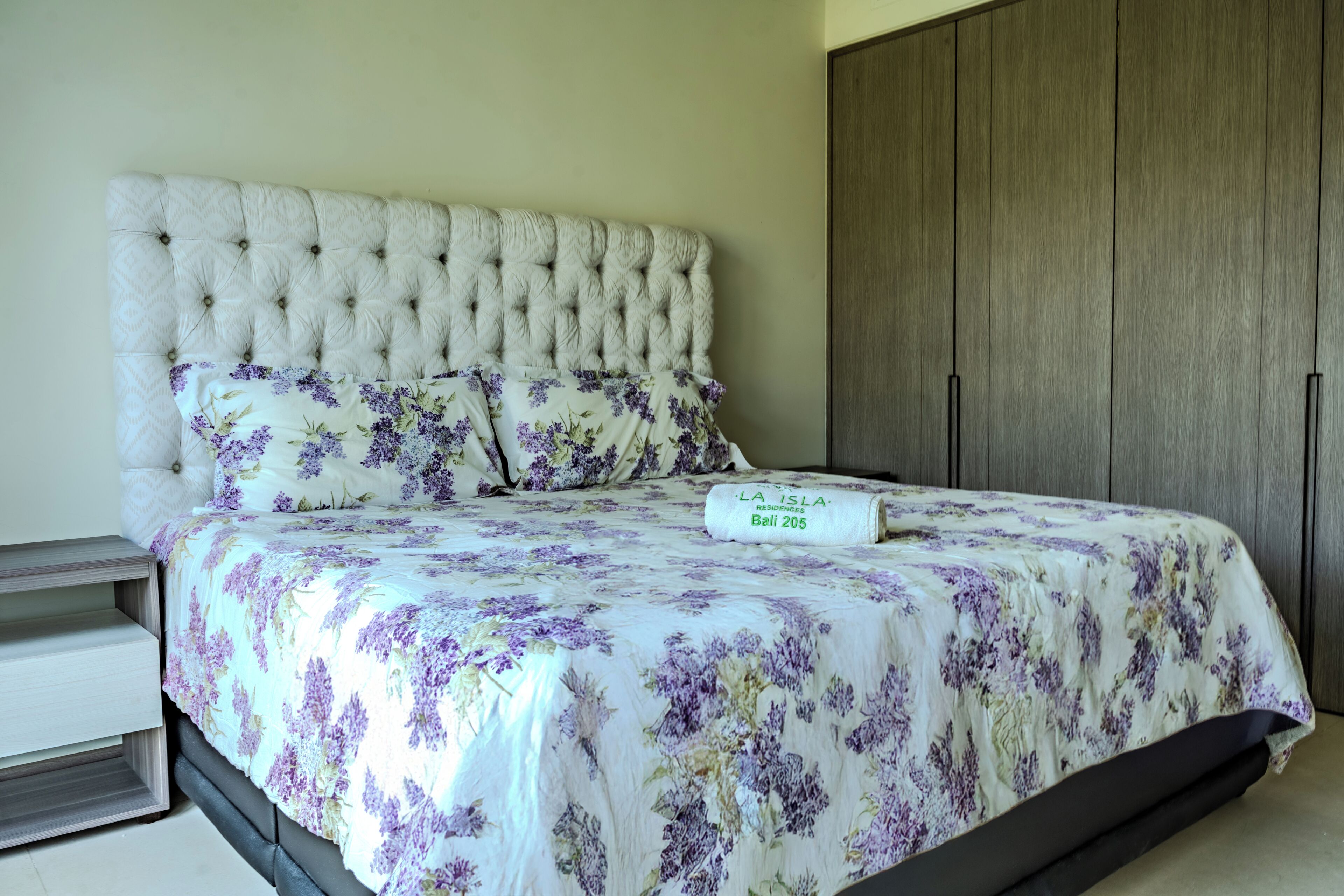 Un dormitorio con cabecera capitonada, ropa de cama estampada, mesita de noche y armario.
