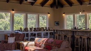Un interior rústico con un alto techo de madera, un rincón acogedor con sofá y sillones, una zona de bar con taburetes y ventanas amplias que ofrecen vistas a la vegetación.