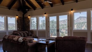 Una sala de estar acogedora con pared de piedra, techo de madera y ventanas amplias con vistas a las colinas.