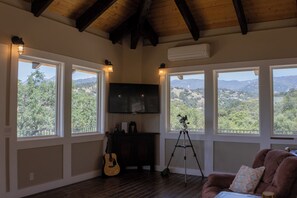 Een kamer met een houten plafond, een tv aan de muur, een statief, een gitaar en een stoel met kussen.