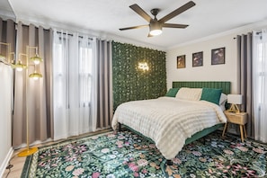 Ein Schlafzimmer mit Bett, Vorhängen, Deckenventilator, Wandleuchte und einem blumigen Teppich.