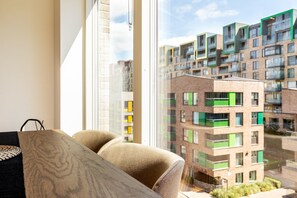 Ein modernes Apartment mit großem Fenster, einer Couch und Blick auf ein mehrstöckiges Gebäude mit Balkonen.