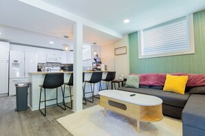 Eine moderne Küche mit weißen Schränken, einer Bar inklusive Hocker, einem Esstisch sowie einem Wohnbereich mit einer Couch und Kissen.